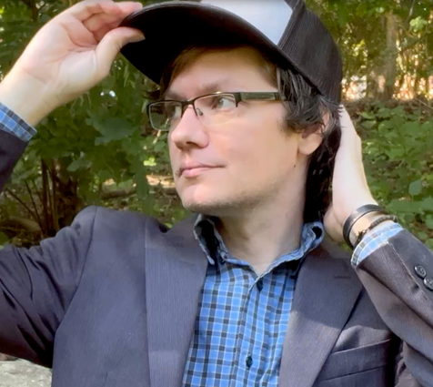 Jared the freelancer. He is a male wearing glasses, a ball cap, and a suit jacket, and bracelets. He is looking to the left.